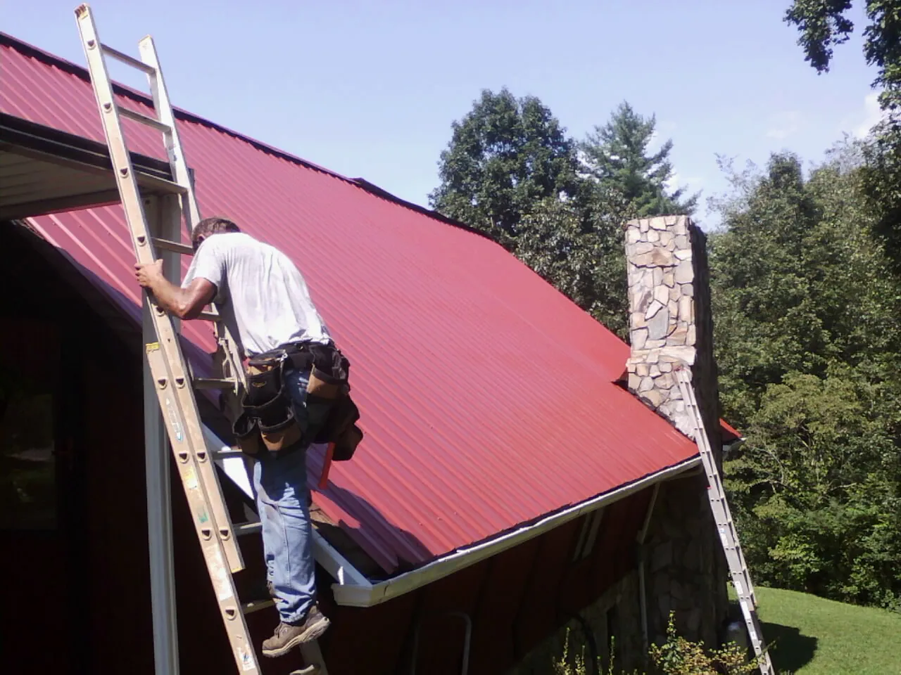 Roofer on a ladder beside a red metal roof and stone chimney — Soffit & Fascia Repair by Roofing Brothers in Hallieford